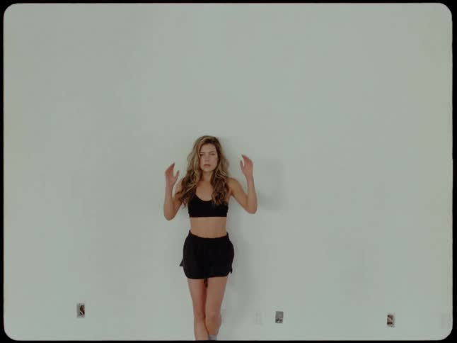 A beautiful woman with long wavy hair opens her eyes and gazes at the camera as it slowly zooms out to reveal her posing against a white wall
