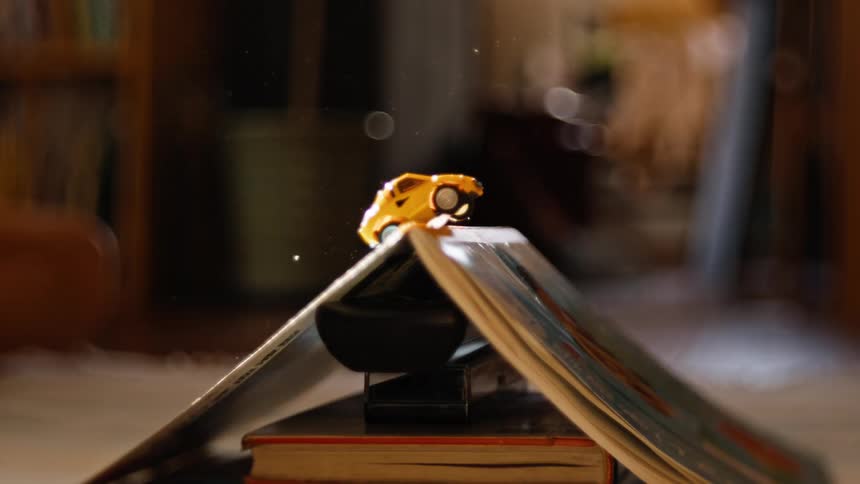 A yellow toy car drives over a book ramp in a whimsical slow-motion close-up as dust particles float in the air