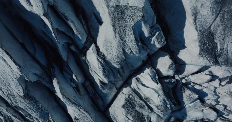 An abstract top-down aerial view slowly pulls back to reveal the immense scale and intricate, shadowy textures of a glacier