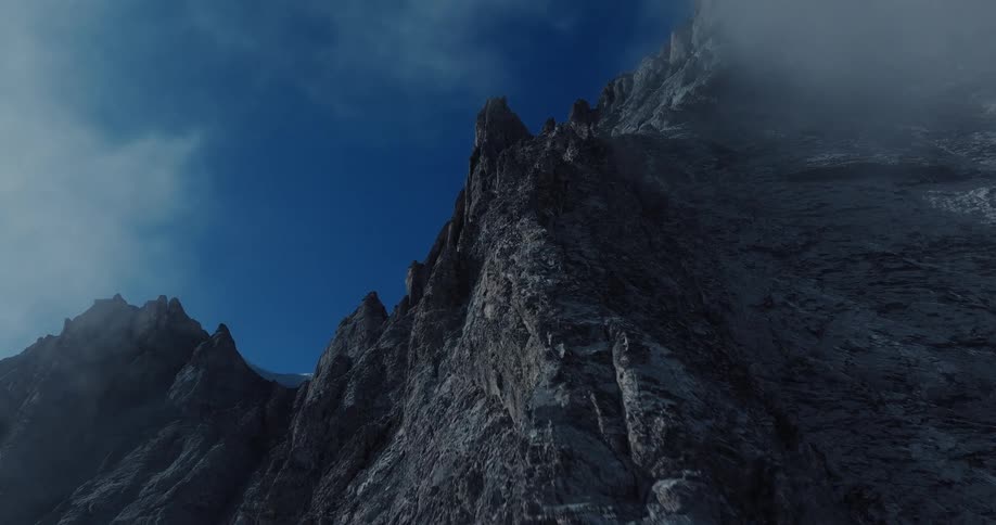 A slow, dramatic CG animation climbs up the face of a vast, jagged mountain peak as clouds and fog drift by against a deep blue sky