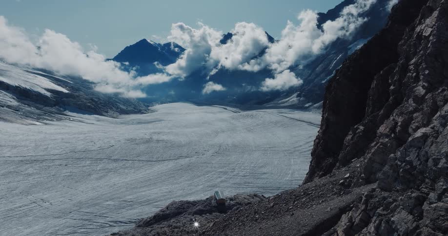 A stunning wide shot captures a vast glacier valley nestled between towering, cloud-covered mountain peaks