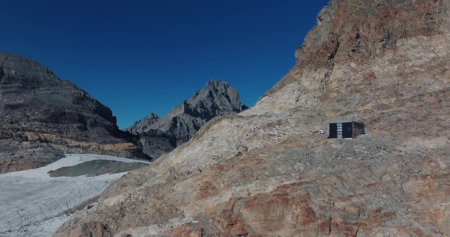 An isolated, modern cabin sits on a rocky mountain slope overlooking a vast glacier in a high alpine environment