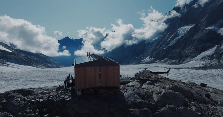 An epic view of a remote mountain refuge and helicopter perched on a rocky outcrop amidst a sprawling snow-covered glacier in the Alps
