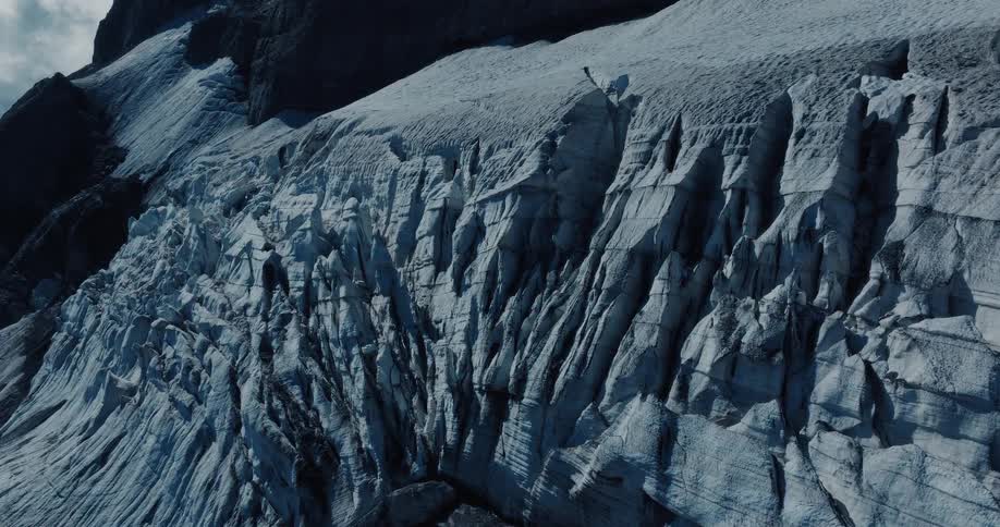 A static shot focuses on the deeply crevassed and textured face of a massive glacier on a dark, rocky mountain