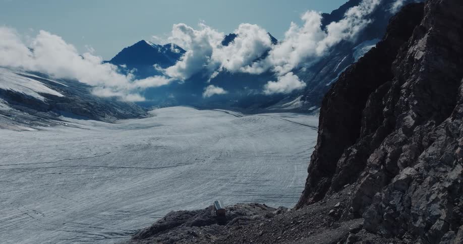 A stunning, static view of a massive glacier flowing through a valley surrounded by majestic, cloud-covered mountains