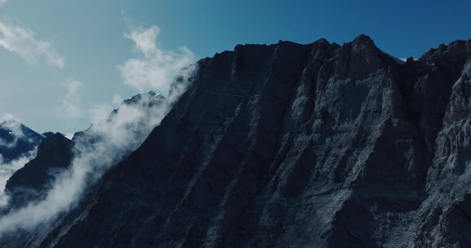 A dramatic close-up of a dark, rocky mountain peak with clouds rolling over its ridges against a clear blue sky