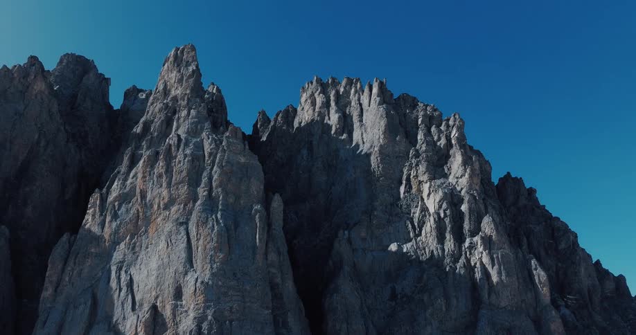 A slow, majestic aerial shot reveals the jagged, rocky peaks of a mountain range against a clear blue sky