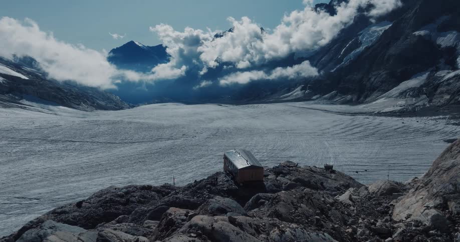 A static, wide shot shows a small, isolated cabin on a rocky cliff overlooking a vast glacier in a dramatic mountain valley
