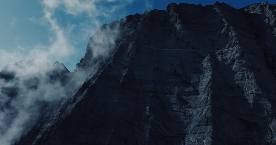 A dramatic low-angle shot of a massive shadowed mountain with clouds drifting across the deep blue sky