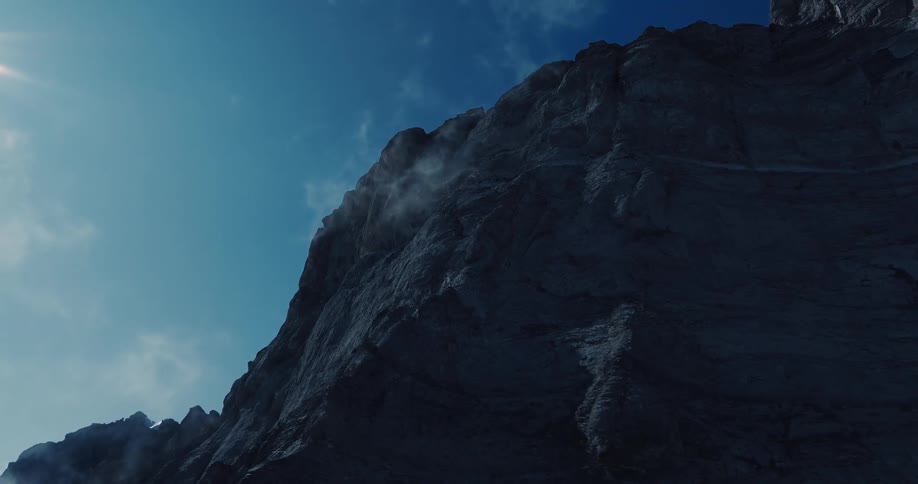 An imposing rocky mountain face looms in shadow as mist drifts from its peak under the bright sun