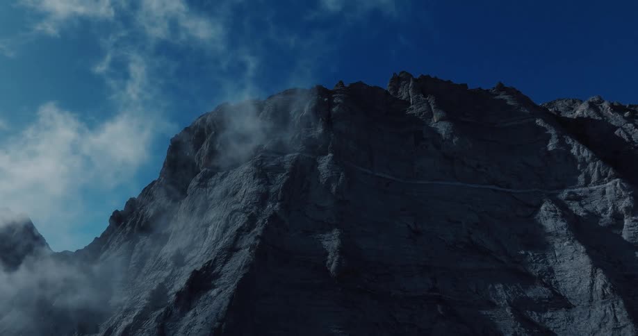 Wispy clouds drift down the shadowy face of a rugged mountain peak, revealing a narrow trail against a deep blue sky