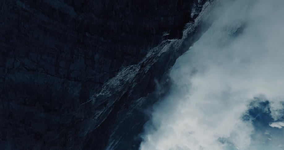 A vertical shot of a powerful waterfall cascading down a dark, rocky mountain cliff face with clouds swirling below