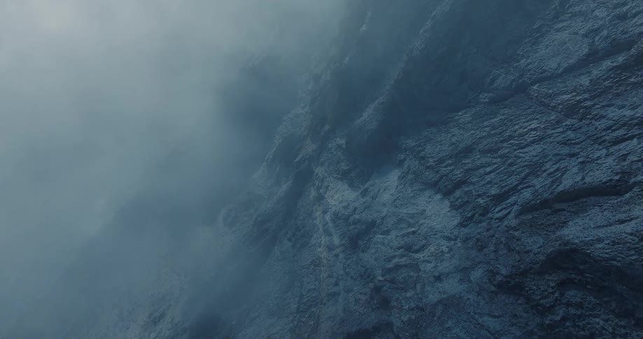 A vertical view of a vast, rocky cliff face shrouded in swirling fog