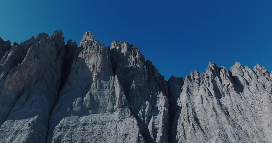 A static low-angle shot captures the rugged, rocky peaks of a mountain range against a cloudless, deep blue sky