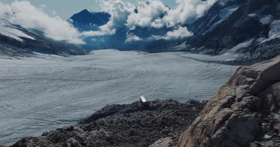 A solitary, modern cabin perches on a rocky cliff overlooking a vast glacier in a mountain valley under a cloudy sky
