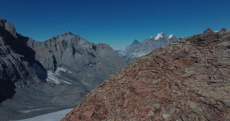 An aerial shot pans over a rocky mountain ridge, revealing a breathtaking view of a glacial valley and snow-capped peaks in the Alps