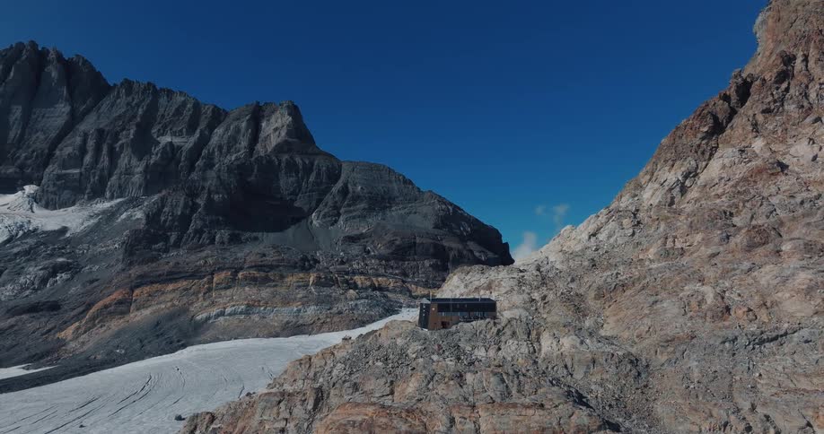 An isolated modern cabin sits on a rocky cliffside in a vast mountain range next to a glacier on a clear day