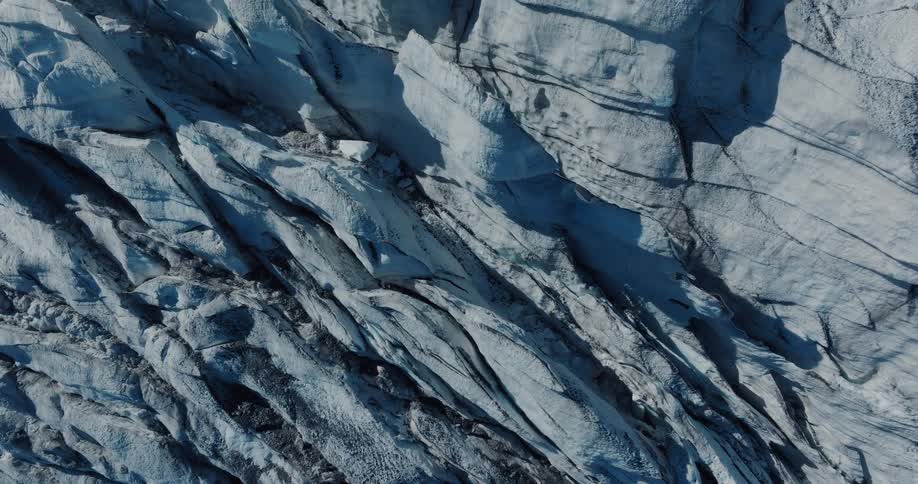 An abstract top-down aerial shot reveals the dramatic textures and deep blue cracks of a vast ice glacier