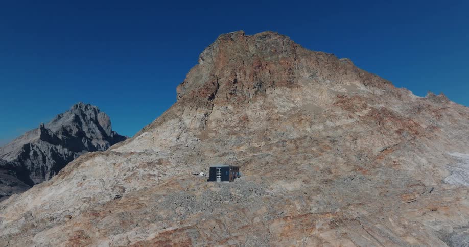 Aerial view of a remote mountain hut perched on a vast rocky peak next to a glacier under a clear blue sky