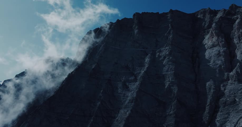 A dramatic low-angle shot of a shadowy mountain peak with clouds swirling around its steep rock face under a blue sky