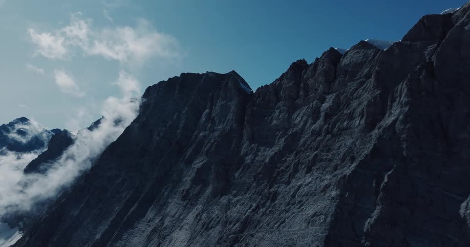 Epic aerial footage slowly approaches a towering, rugged mountain peak partially shrouded in clouds