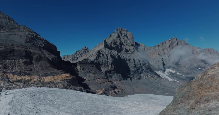 Slow aerial flight over a massive glacier nestled between towering, jagged mountain peaks under a clear blue sky