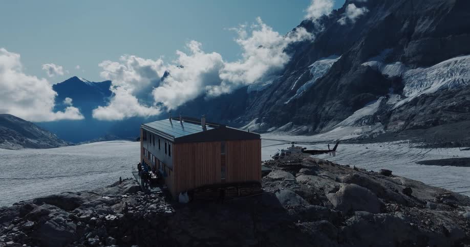 A majestic view of a remote mountain hut on a rocky ledge with a landed helicopter, overlooking a vast glacier surrounded by snowy peaks