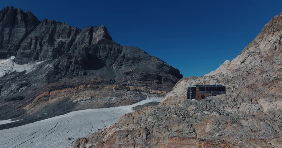 A majestic slow aerial pullback reveals a modern mountain hut nestled on a rocky cliff overlooking a vast glacier in the Alps
