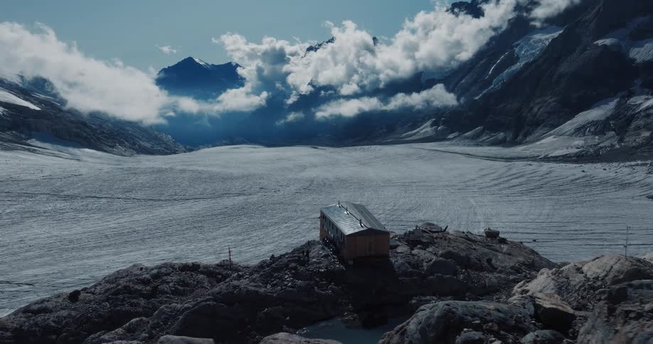 A solitary wooden cabin sits on a rocky cliff overlooking a vast glacier in a dramatic, cloud-filled mountain landscape