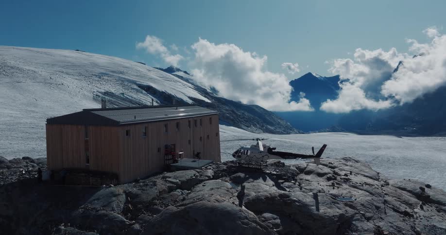 A helicopter is parked next to a modern wooden mountain refuge on a rocky outcrop, surrounded by a vast glacier and snow-covered peaks