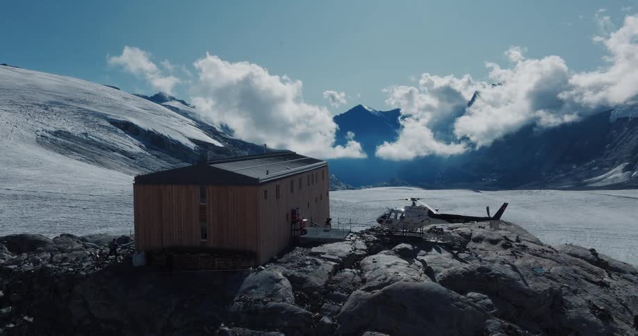 A modern wooden mountain refuge with a parked helicopter overlooks a vast glacier in the Swiss Alps