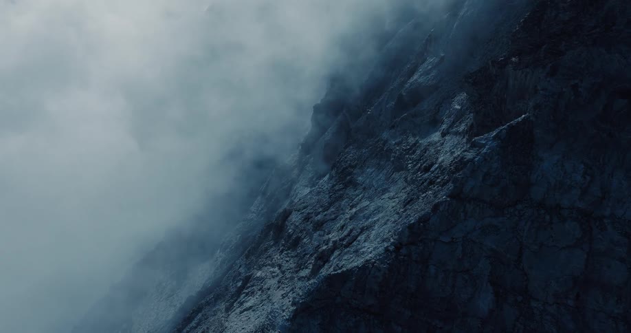 A mysterious and cold mountain face emerges from the dense, swirling fog on a dramatic day