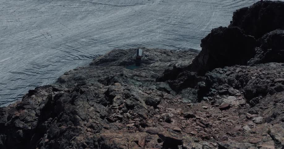 A solitary mountain shelter sits on a rugged rock cliff overlooking a vast, icy glacier in a desolate landscape