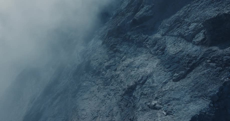 A slow and moody vertical aerial shot pushes in on a dark, rocky mountain face shrouded in thick fog