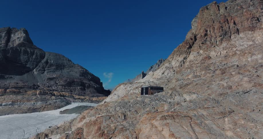 A solitary mountain refuge stands on a rocky outcrop above a glacier in the Swiss Alps on a clear day