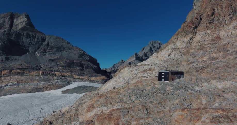 A slow pan reveals a modern, isolated alpine hut nestled on a rocky mountainside overlooking a massive glacier under a clear blue sky