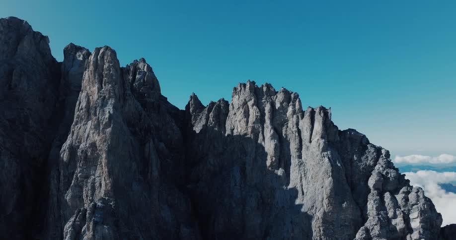 A stunning aerial view reveals a jagged mountain peak piercing a clear blue sky, with a sea of clouds visible in the distance