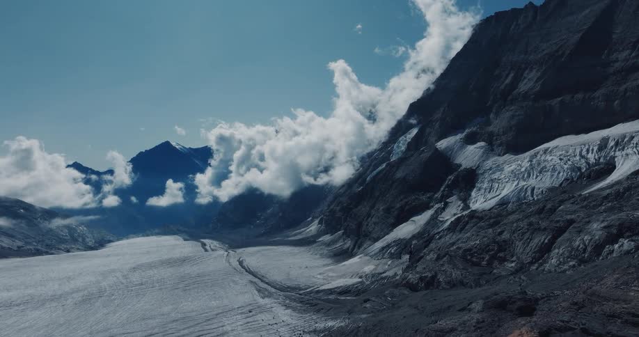 An epic aerial drone shot slowly flies over a vast glacier in a mountain valley under a blue, cloudy sky