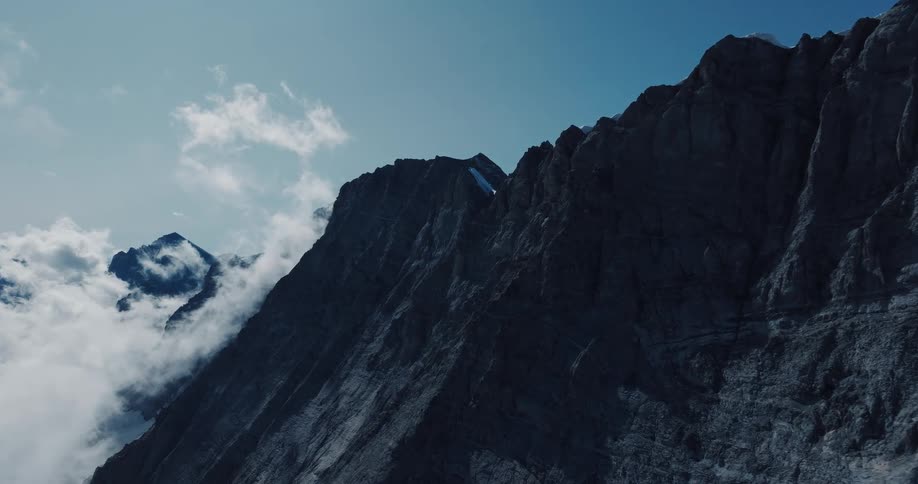 A majestic and imposing rocky mountain peak looms against a clear blue sky as soft clouds drift by