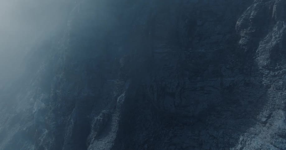 Dark, moody fog drifts slowly across the textured face of a rugged cliff
