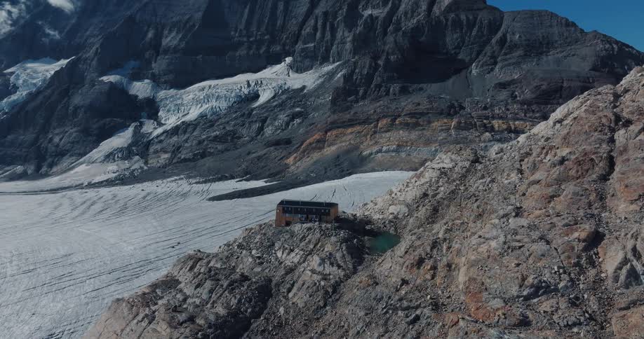 A solitary hut stands perched on a rocky cliff overlooking a vast glacier nestled within a majestic mountain range