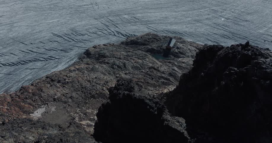 High angle aerial shot reveals a solitary modern hut nestled on a rocky mountain peak next to a sprawling, textured glacier