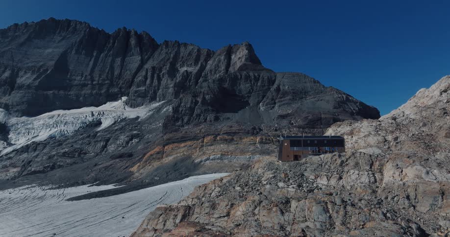 A stunning aerial view reveals a remote and modern mountain hut nestled in a vast landscape of rocky peaks and a sprawling glacier