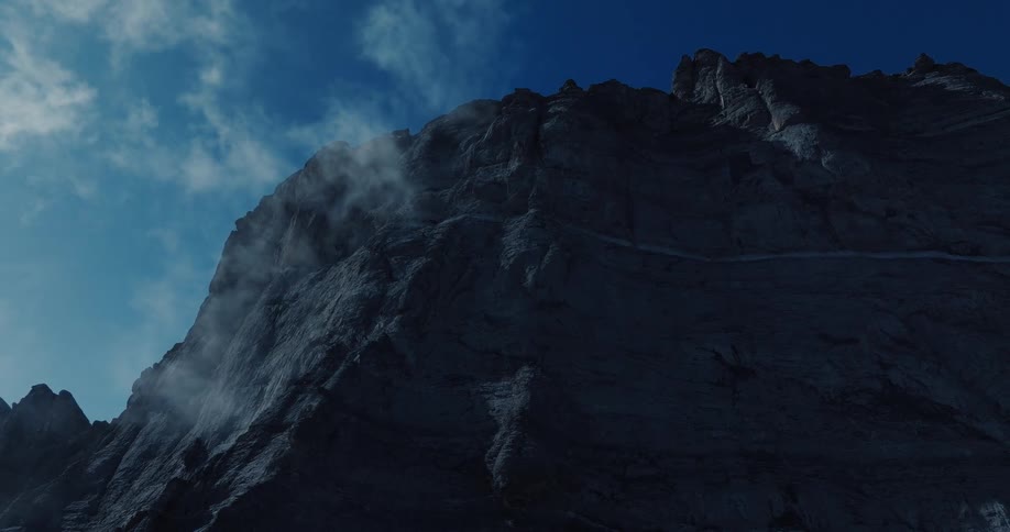 A dramatic low-angle reveal of a massive, shadowy mountain face with mist swirling around its peaks against a deep blue sky