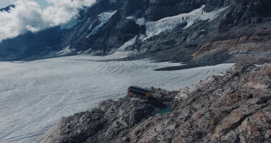 A slow aerial zoom-in reveals a remote mountain hut perched on a rocky cliff overlooking a vast glacier