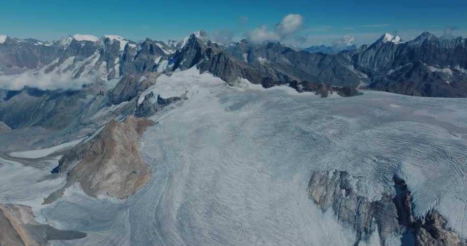 A majestic aerial panorama showcases a vast glacier surrounded by towering, snow-capped mountain peaks under a brilliant blue sky