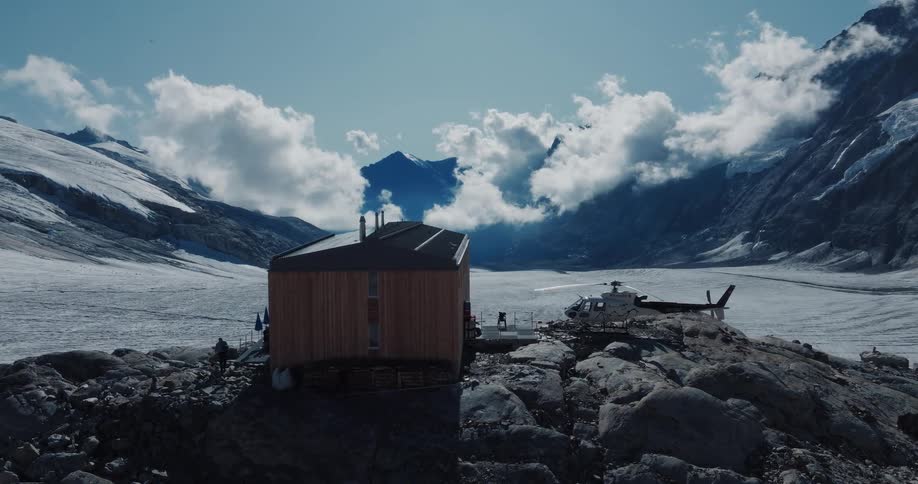 A helicopter sits next to a remote wooden hut on a rocky peak, surrounded by a breathtaking glacier and snow-capped mountains