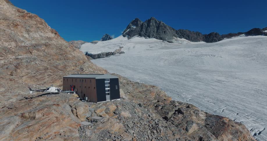 Aerial pull-back shot reveals a modern alpine hut with a helicopter parked next to a vast glacier in the mountains