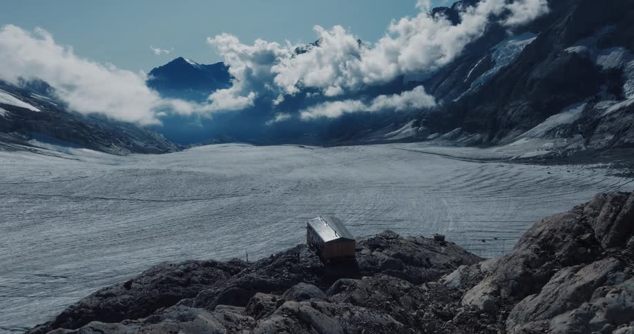 A small remote cabin sits on a rocky outcrop overlooking a vast glacier surrounded by majestic mountains