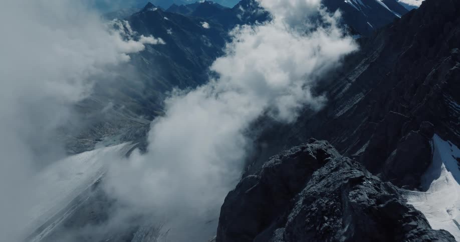 An epic aerial shot pushes in and tilts down over a rugged, snow-dusted mountain peak, revealing the vast, misty valley floor below